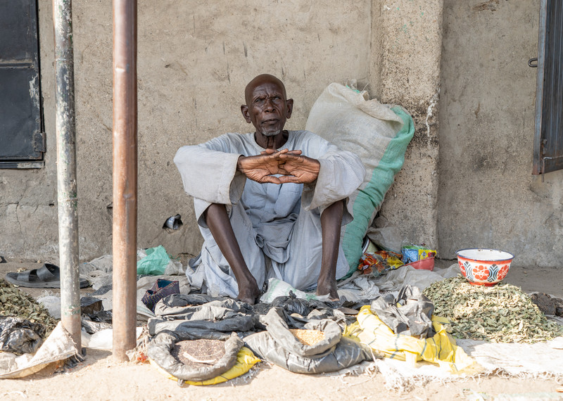 在奈及利亞東北部博爾諾州（Borno State）邁杜古里（Maiduguri）附近的市場上，一位老人坐著等待出售食物。© The Walking Paradox / Amnesty International